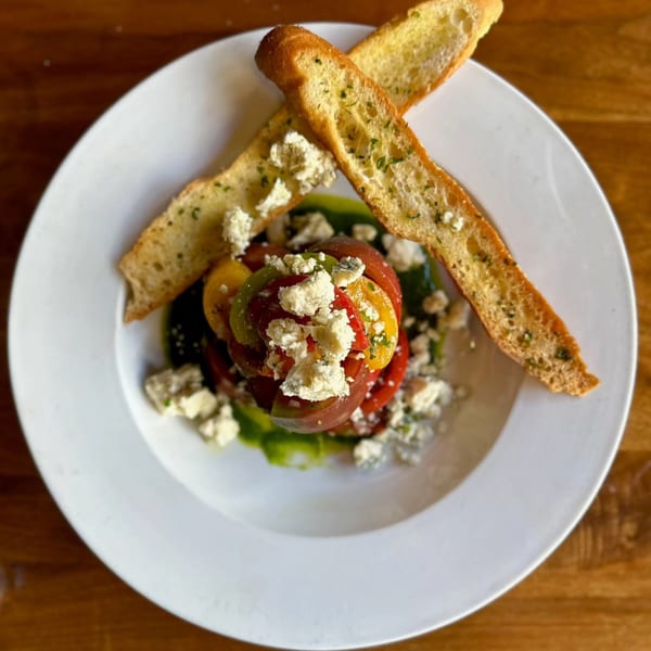 Heirloom tomato salad with crumbled feta cheese, green sauce, and two garlic bread slices on a white plate.