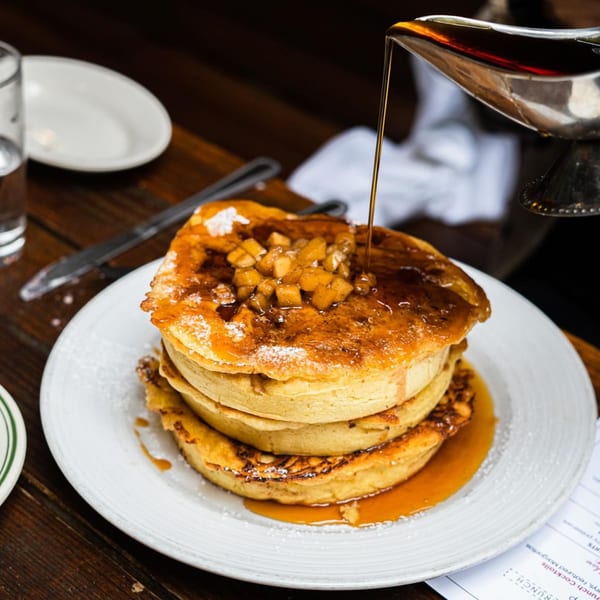 Maple syrup pouring over a stack of pancakes topped with diced apples and powdered sugar.