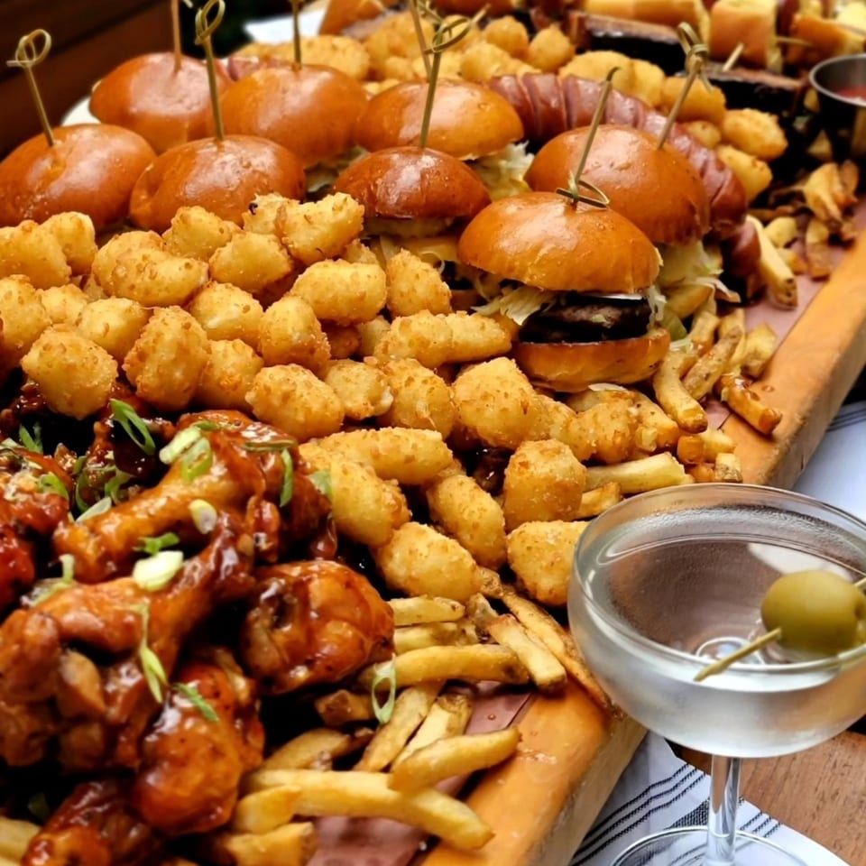 A wooden board piled with sliders, tater tots, french fries, chicken wings, and a martini.