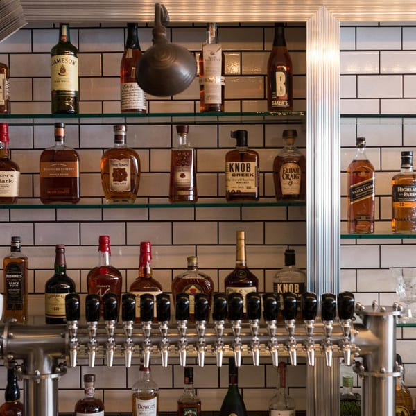 A well-stocked bar at Butcher Bar with various liquor bottles and beer taps against a white tiled wall.