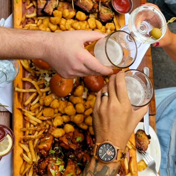 Four people toast drinks over a large Butcher Bar platter of fries, tots, wings, and sliders.